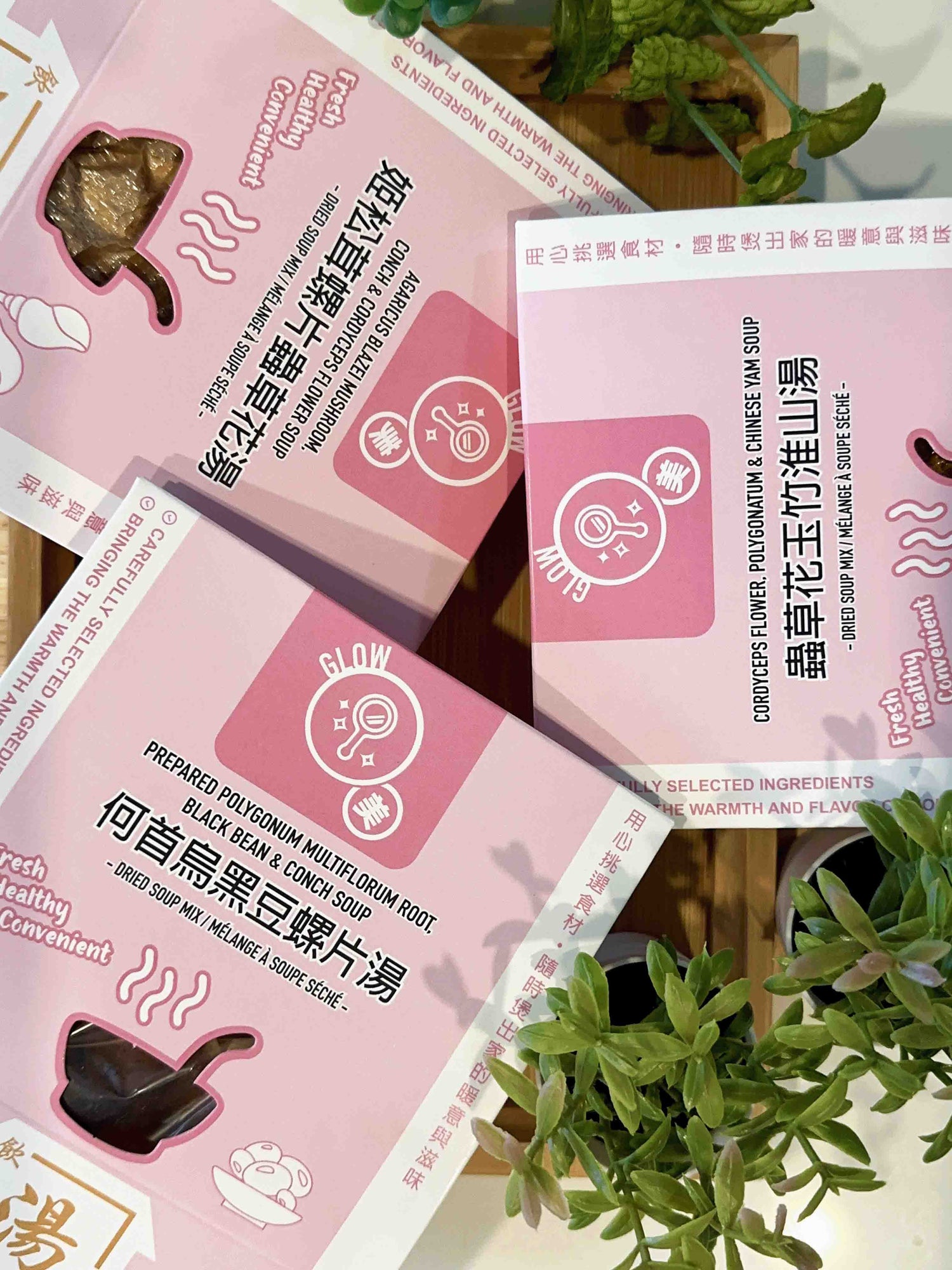 Glow Series herbal soup packs — Cordyceps Flower Soup, Polygonatum & Chinese Yam Soup, and Prepared Polygonum Multiflorum Root Soup displayed on a wooden table with greenery. 飲湯思源 GLOW 系列湯包，包括蟲草花玉竹淮山湯、姬松茸螺片蟲草花湯、何首烏黑豆螺片湯，木桌佈景搭配綠植，自然健康感。