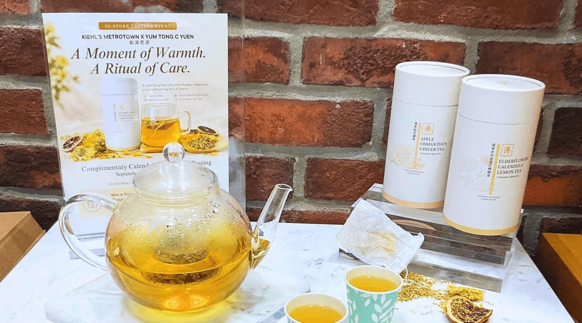 In-store display for the Kiehl's Metrotown x Yum Tong C Yuen tasting event — a glass teapot of brewed Elderflower Calendula Lemon Tea, two tea canisters, and the event poster against a brick wall. Kiehl's Metrotown 與飲湯思源合作試飲活動嘅店內陳設——玻璃茶壺沖泡住接骨木花金盞花檸檬茶，配兩罐茶葉同活動海報，背景係磚牆。