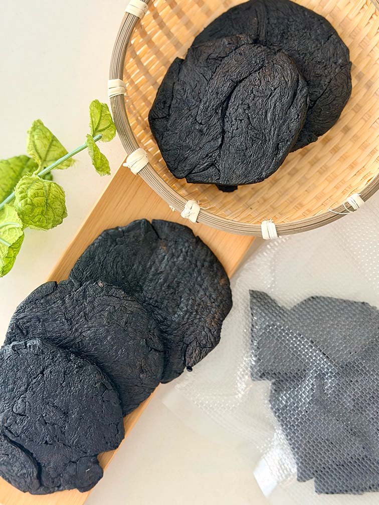 Shu Di Huang Prepared Rehmannia Root pieces in a bamboo basket beside vacuum-sealed packaging 熟地炮製地黃根塊置於竹籃中，旁邊展示真空密封包裝