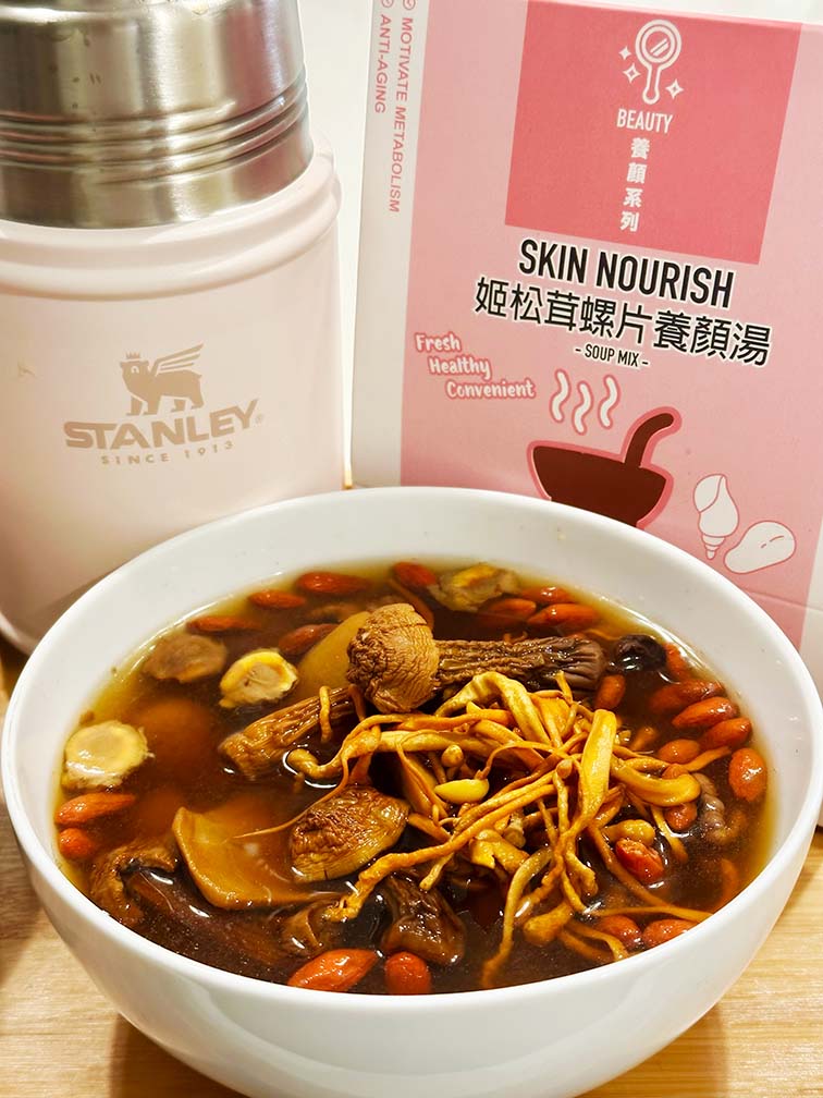 A bowl of warm soup accompanied by a Stanley food jar in Rose Quartz and a skin nourish soup pack on a wooden table, creating a cozy dining atmosphere. 木桌上一碗熱騰騰的暖湯，配上玫瑰粉色Stanley燜燒壺和姬松茸螺片養顏湯包，營造出溫馨的用餐氛圍。