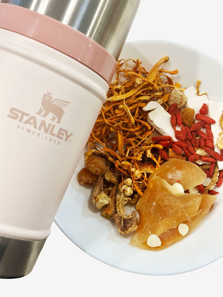 A bowl filled with skin nourish soup's ingredients and Chinese herbs placed next to a Stanley food jar in Rose Quartz on a wooden surface. 一個裝滿姬松茸螺片養顏湯湯料食材的碗放在木質表面上的玫瑰粉色Stanley 燜燒壺旁