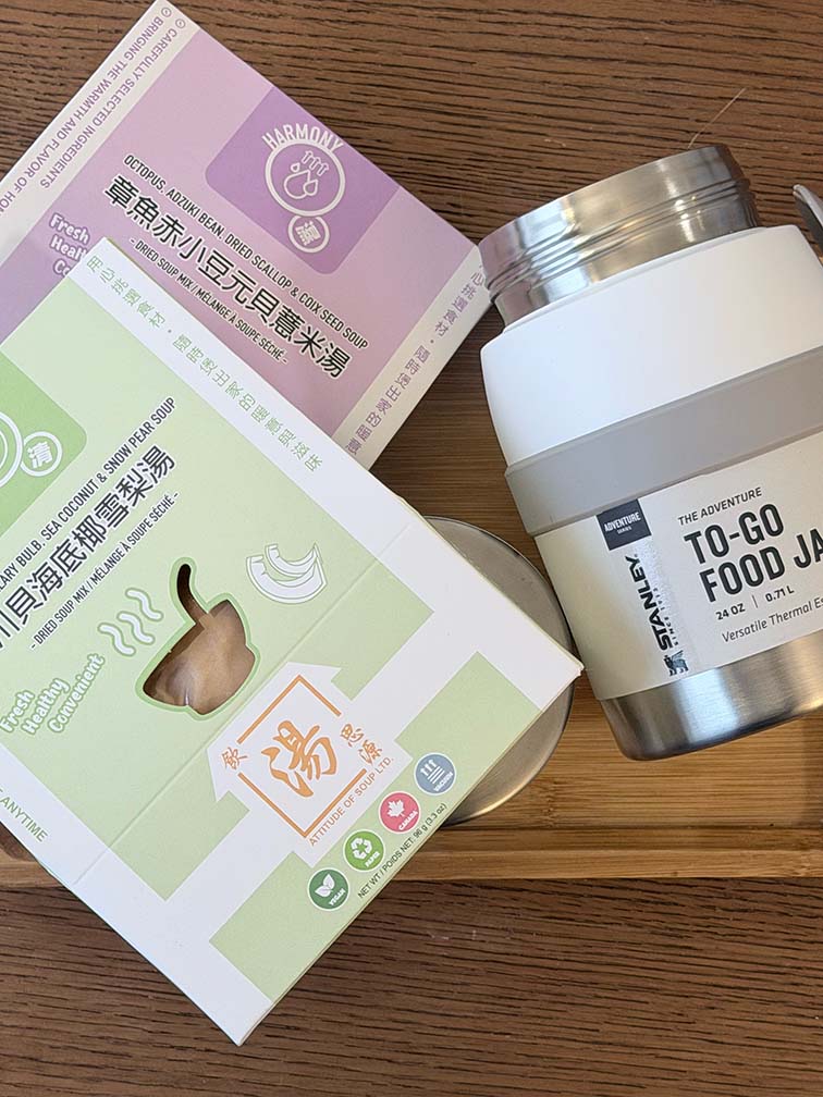Angled top-down photo on a wooden surface showing two boxed Chinese soup mixes (Octopus, Adzuki Bean, Dried Scallop & Coix Seed Soup and Sichuan Fritillary Bulb, Sea Coconut & Snow Pear Soup) on the left and a white Stanley Adventure To-Go Food Jar on the right with the lid removed, exposing the stainless-steel threads. 木質桌面斜俯視照片：左側有兩盒湯包 (川貝海底椰雪梨湯、章魚赤小豆元貝薏米湯），右側是一個白色 Stanley Adventure To-Go Food Jar，罐蓋已打開，露出不鏽鋼螺紋口。
