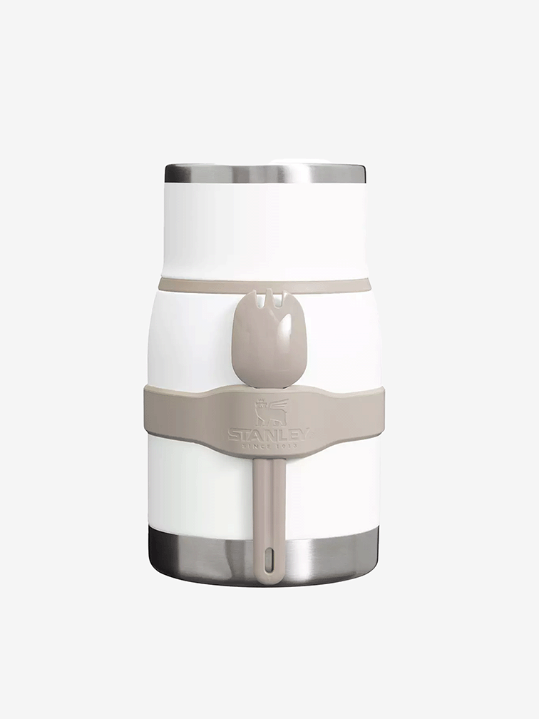 Front view of a Stanley Adventure To-Go white insulated food jar with silver metal rim and base, wrapped with a beige band holding a matching spoon. 白色背景上的Stanley Adventure To-Go 白色保溫食物罐正面照，上下為銀色金屬邊與底座；中間有米色固定帶並夾著同色湯匙，固定帶上有壓印的 Stanley 字樣。