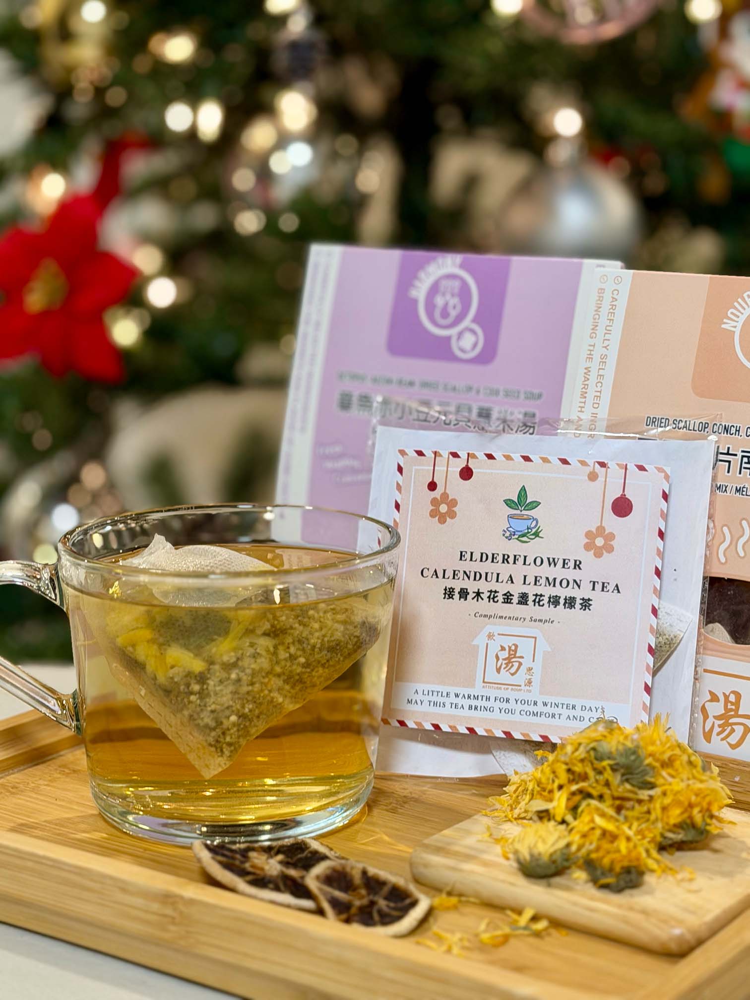 A glass mug of steeping elderflower calendula lemon tea beside dried ingredients and Christmas décor, featuring Yum Tong Cyuen’s complimentary holiday flower tea sample packaging. 玻璃杯沖泡中的接骨木花金盞花檸檬花茶，旁邊放有乾燥材料與聖誕裝飾，呈現飲湯思源節日限定花茶試飲包。